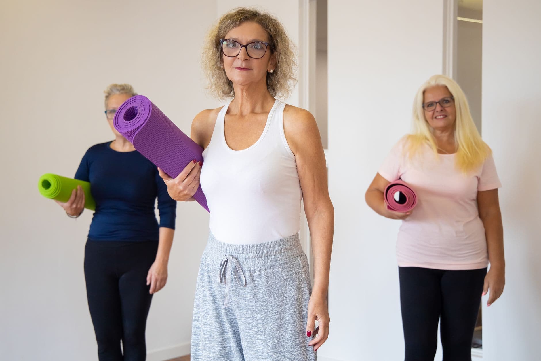 três mulheres de mais idade segurando tapetes de exercício antes de uma aula